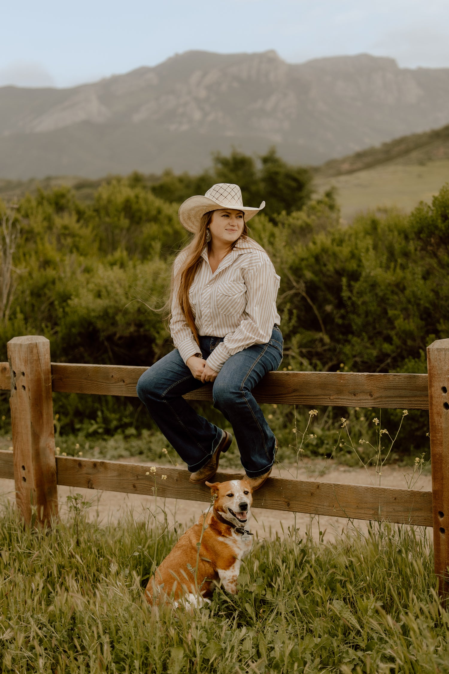 Woman in a cowboy hat sitting on a wooden fence with a dog in a natural setting